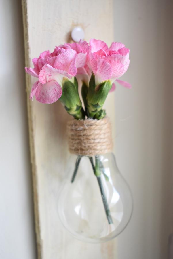 Ręcznie robiony wazon z żarówki obwiązany jutowym sznurkiem, przymocowany do drewnianej deski, z różowymi goździkami. Widoczny biały pinezka.