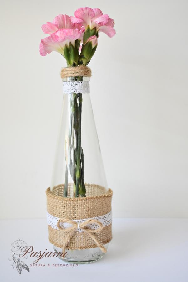 Dekoracyjna butelka z różowymi goździkami, ozdobiona jutą, koronką i sznurkiem, styl rustykalny.