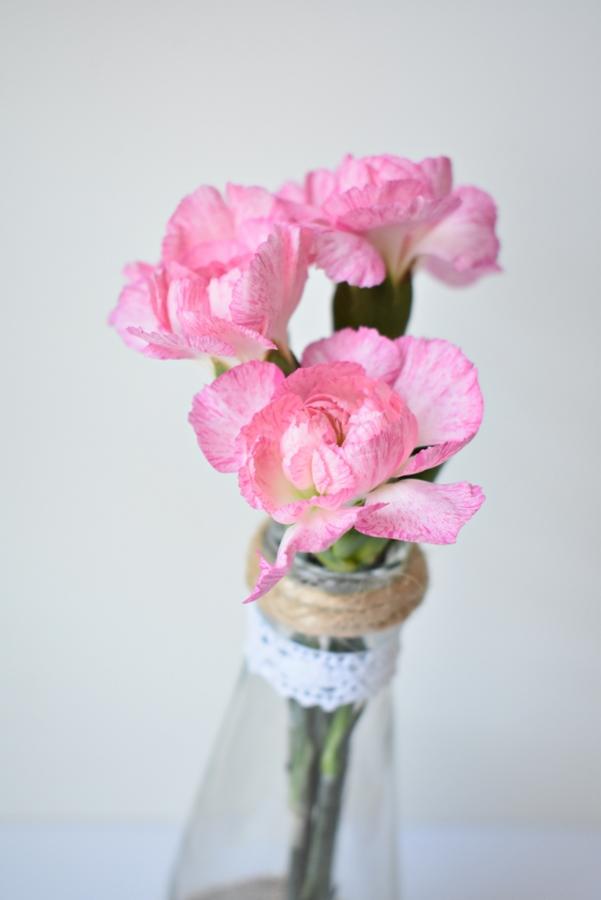 Trzy różowe goździki w szklanej butelce ozdobionej jutowym sznurkiem i białą koronką na jasnym tle.