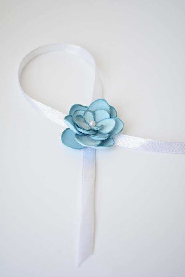 Dekoracyjna, błękitna, materiałowa róża z perłą w środku, umieszczona na białej satynowej wstążce na białym tle.