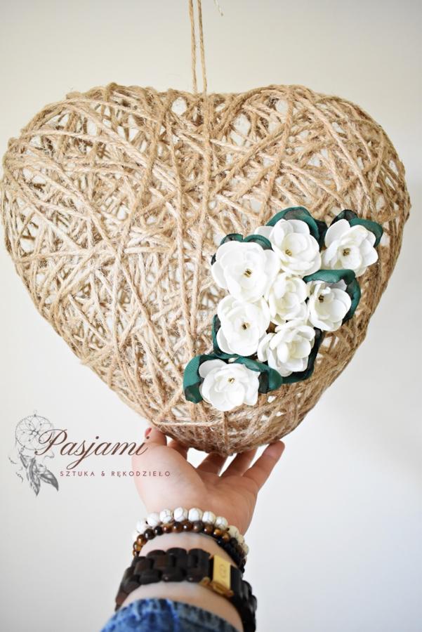 Ręcznie robiona dekoracja w kształcie serca, owinięta jutowym sznurkiem i ozdobiona białymi kwiatami z zielonymi liśćmi, trzymana w dłoni z widocznymi bransoletkami.