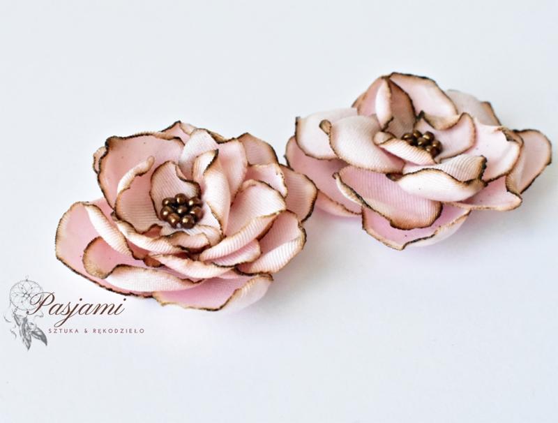 Dwa ręcznie robione, różowe kwiaty z materiału z brązowymi krawędziami i środkiem, leżące na białym tle, z widocznym logo 'Pasjami Sztuka & Rękodzieło'.