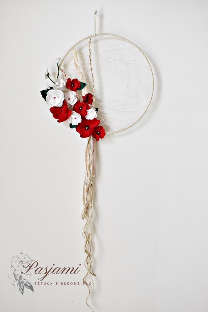 Okrągły łapacz snów z białymi i czerwonymi kwiatami z filcu, z beżowymi sznurkami zwisającymi na białym tle ściany, z widocznym logo firmy w lewym dolnym rogu.