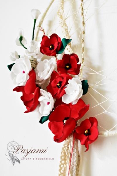 Ręcznie robiony łapacz snów z białymi i czerwonymi kwiatami z filcu, zawieszony na białej ścianie, z widocznym logo firmy w lewym dolnym rogu.