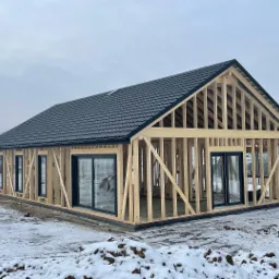 Surowa konstrukcja domu z drewnianym szkieletem, częściowo pokryta czarną dachówką, widoczne czarne ramy okienne, zasypana śniegiem.