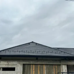 Surowy stan budynku z ciemną dachówką na tle pochmurnego nieba, widoczne elementy konstrukcyjne i okna