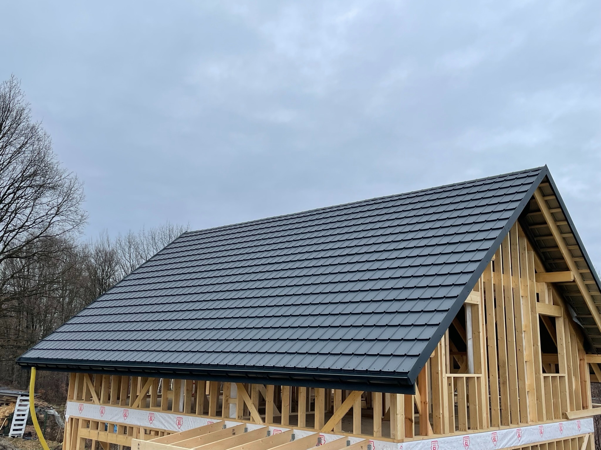 Dom w trakcie budowy, widoczna drewniana konstrukcja szkieletowa i pokryty grafitową blachodachówką dach, z zamontowanymi rynnami, pochmurne niebo w tle.