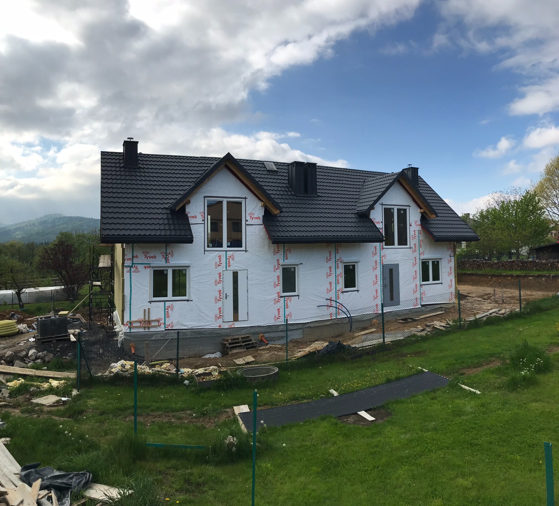 Niedokończony dom z czarnym dachem krytym dachówką, zabezpieczony membraną, widok na wzgórza w tle.