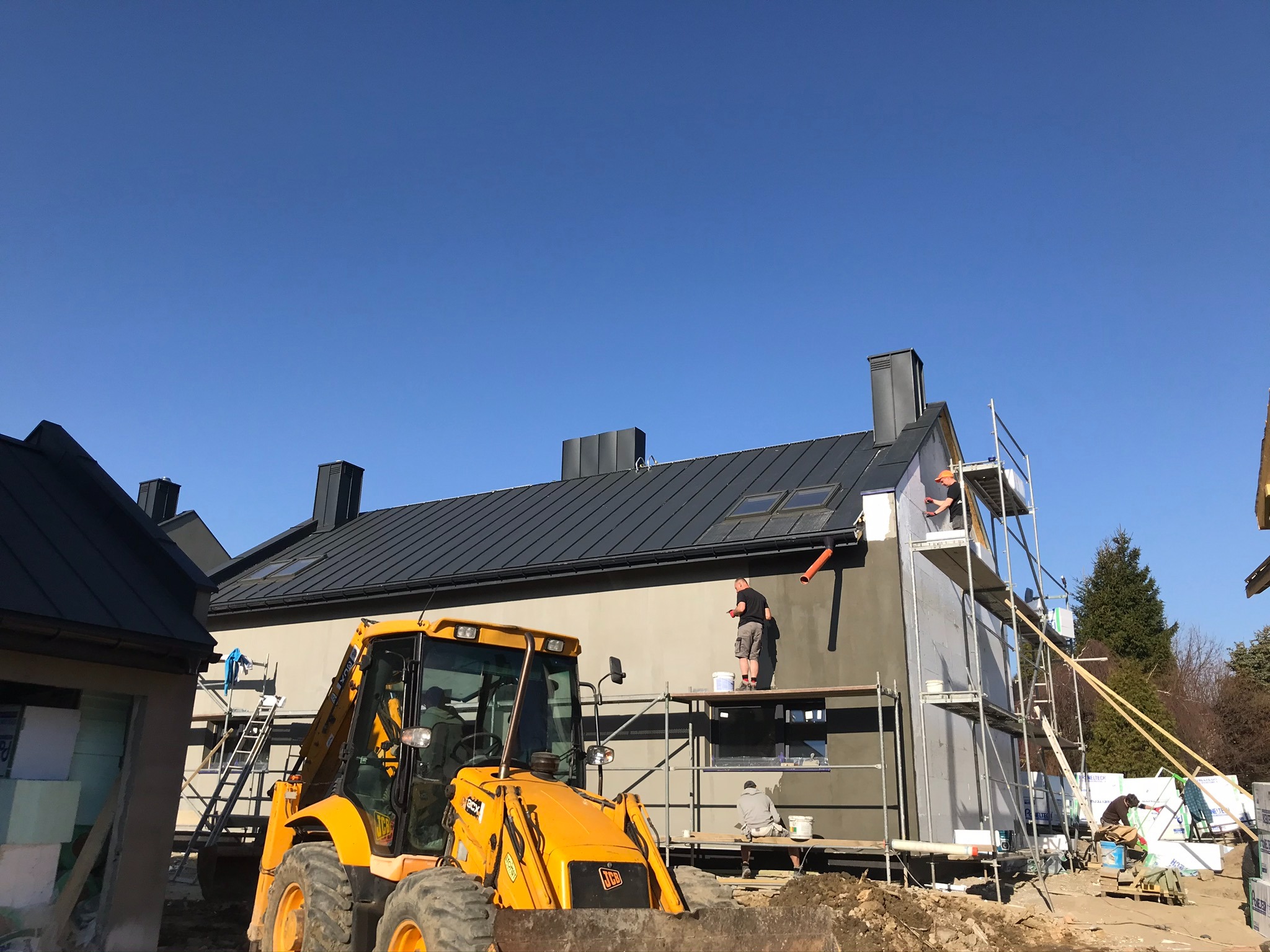 Prace wykończeniowe na elewacji domu szkieletowego z wykorzystaniem rusztowań i żółtej koparki JCB, w tle błękitne niebo.