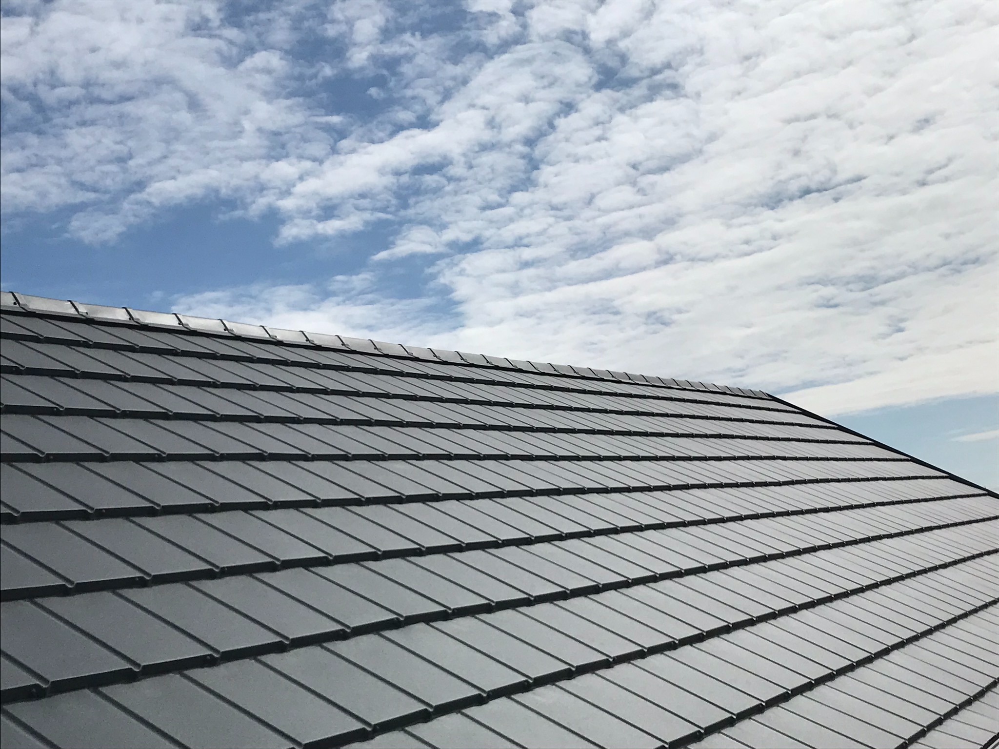 Grafitowe dachówki ceramiczne ułożone pod kątem na tle błękitnego nieba z białymi obłokami.