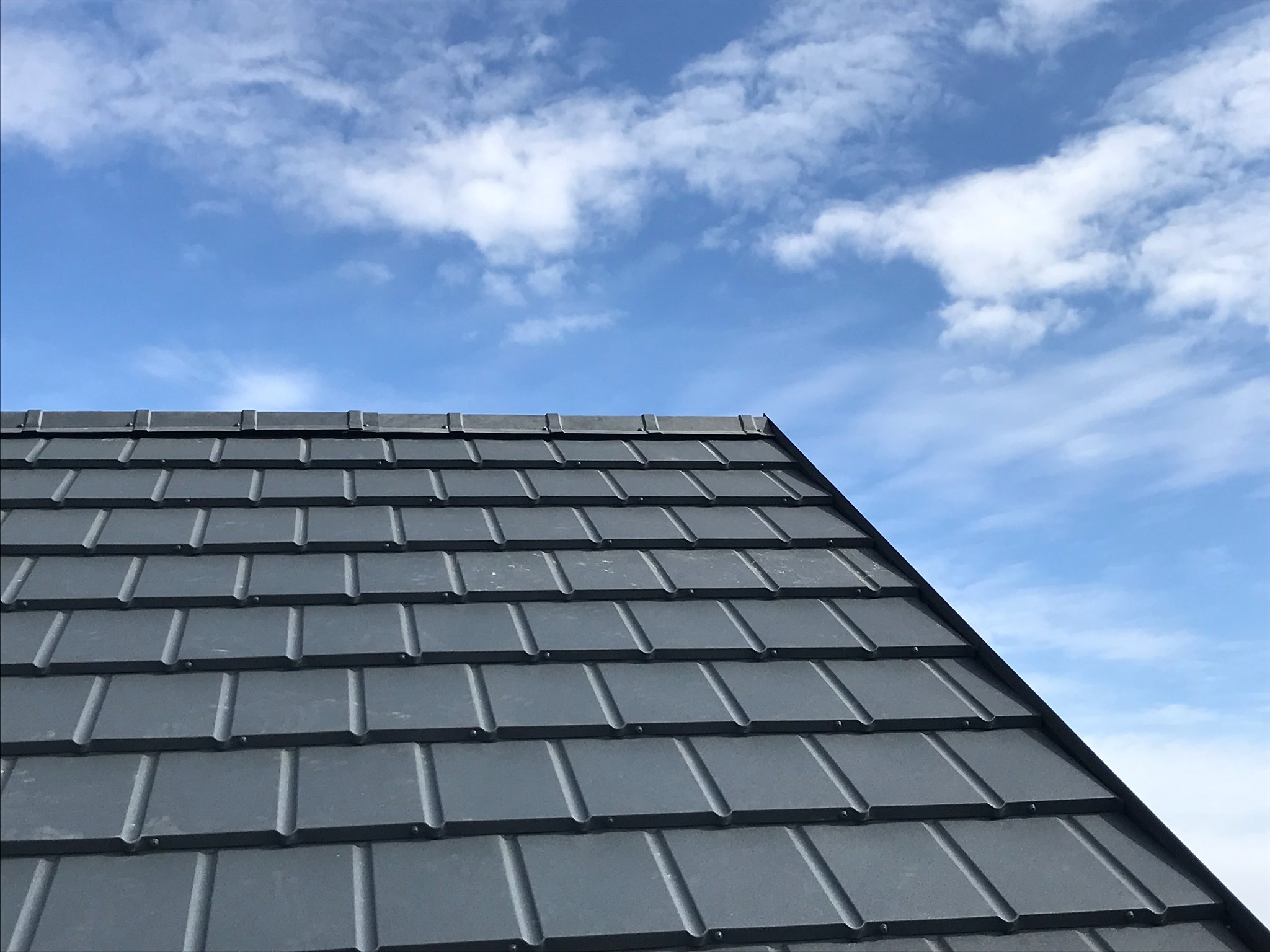 Fragment dachu pokrytego szarymi dachówkami karpiówkami na tle błękitnego nieba z białymi obłokami.