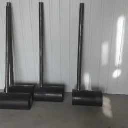 Metalowe konstrukcje spawane z rur i cylindrów, ustawione na szarej podłodze przy białej ścianie z panelami, widoczne ślady spawania.