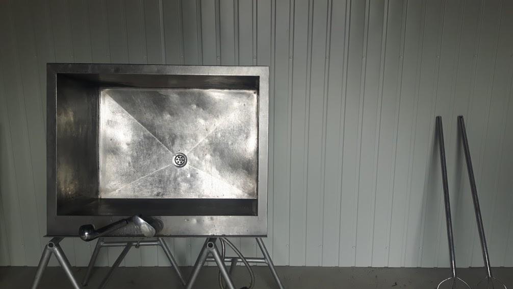 Metalowa wanna na metalowym stelażu z odpływem, obok dwie metalowe tyczki i leżący na wannie metalowy chwytak, na tle blaszanej ściany.