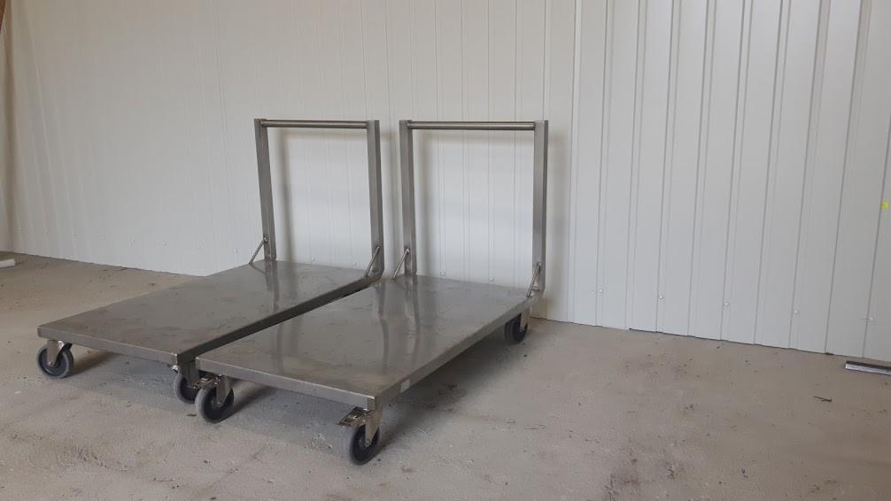 Dwa metalowe wózki transportowe z kółkami, stojące obok siebie w hali z blachy trapezowej. Wózki mają składane uchwyty w kształcie litery U.