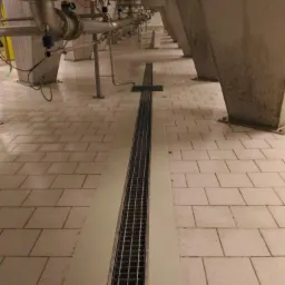 Szczegółowe ujęcie podłogi wyłożonej jasnymi płytkami z centralnie umieszczonym odpływem liniowym ze stali nierdzewnej, poniżej zbiorników na stalowych nogach.