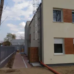 Zewnętrzna drabina ewakuacyjna z osłoną na ścianie budynku mieszkalnego, widoczna z poziomu gruntu, z fragmentem chodnika z kostki brukowej i ogrodzeniem w tle.