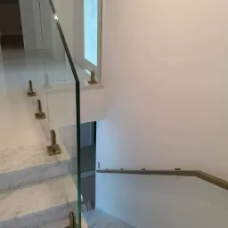 Szklana balustrada na marmurowych schodach z brązowymi mocowaniami, widok z góry na dół.