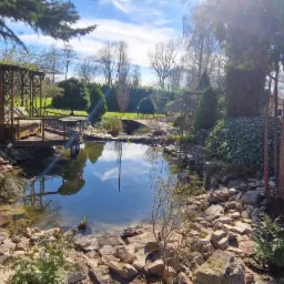 Oczko wodne otoczone kamieniami w zadbanym ogrodzie z drewnianą altaną i mostkiem, w tle widoczne formowane drzewa.