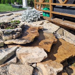 Twój Wymarzony Ogrod - Ogród z kaskadą wodną z kamieni prowadzącą do oczka, obok drewniana altana. Rośliny ozdobne i trawnik w tle. Słoneczny dzień, naturalne materiały.