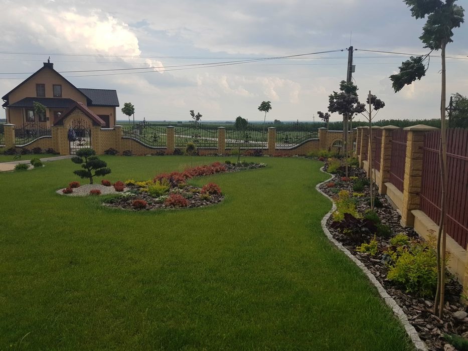 Zadbany trawnik z ozdobnymi rabatami i niską roślinnością wzdłuż ogrodzenia, na tle domu z cegły i rozległych pól uprawnych.
