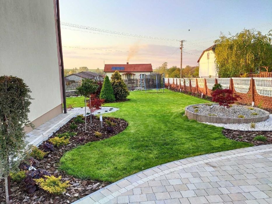 Zadbany ogród przydomowy z równo przystrzyżonym trawnikiem, rabatami z korą i ozdobnymi krzewami, okrągłą rabatą z kamyczkami i małym drzewkiem, widok z perspektywy kostki brukowej.