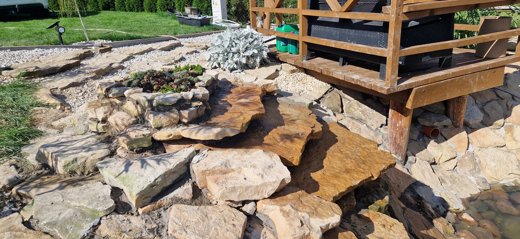 Ogród z kaskadą wodną z kamieni prowadzącą do oczka, obok drewniana altana. Rośliny ozdobne i trawnik w tle. Słoneczny dzień, naturalne materiały.