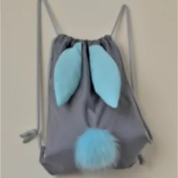 Szary worek-plecak z turkusowymi uszami królika i puszystym pomponem, zawieszony na jasnej ścianie.