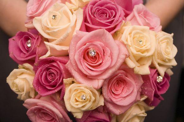 Bukiet róż w odcieniach różu i kremu, ozdobiony drobnymi kryształkami, trzymany w dłoniach na rozmytym, ciemnym tle.