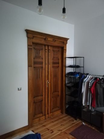 Drewniane, dwuskrzydłowe drzwi wewnętrzne z dekoracyjnymi panelami i ościeżnicą, widoczne w pomieszczeniu z drewnianą podłogą, oświetlone wiszącymi lampami bez kloszy.