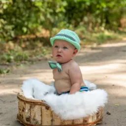 Niemowlę w zielonym, dzierganym kapeluszu i muszce, siedzące w koszyku z brzozowych gałęzi wyłożonym białym futerkiem, na tle zieleni.