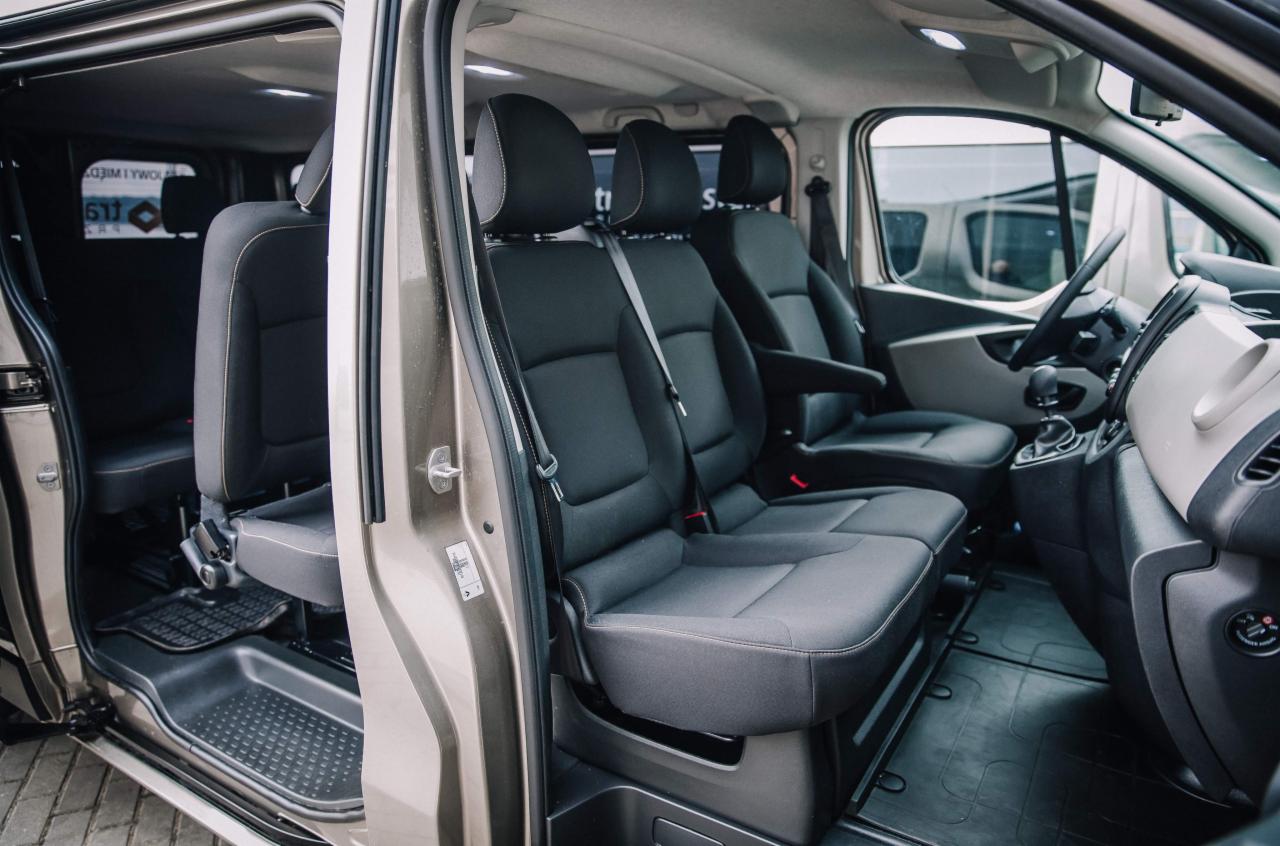 Wnętrze busa z trzema rzędami czarnych foteli, widoczne przez otwarte drzwi, z gumowymi dywanikami na podłodze.