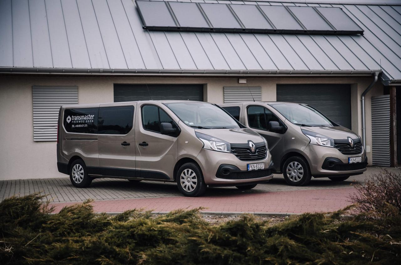 Dwa identyczne, beżowe busy Renault Trafic zaparkowane przed budynkiem z szarym dachem i panelami słonecznymi, z widocznym logo firmy 'transmaster przewóz osób' na oknie jednego z busów.