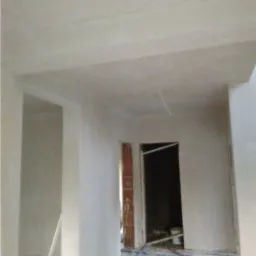 Wnętrze w trakcie prac wykończeniowych: świeżo otynkowane ściany w jasnym odcieniu, widoczne przewody na podłodze i fragment muru w otworze drzwiowym.