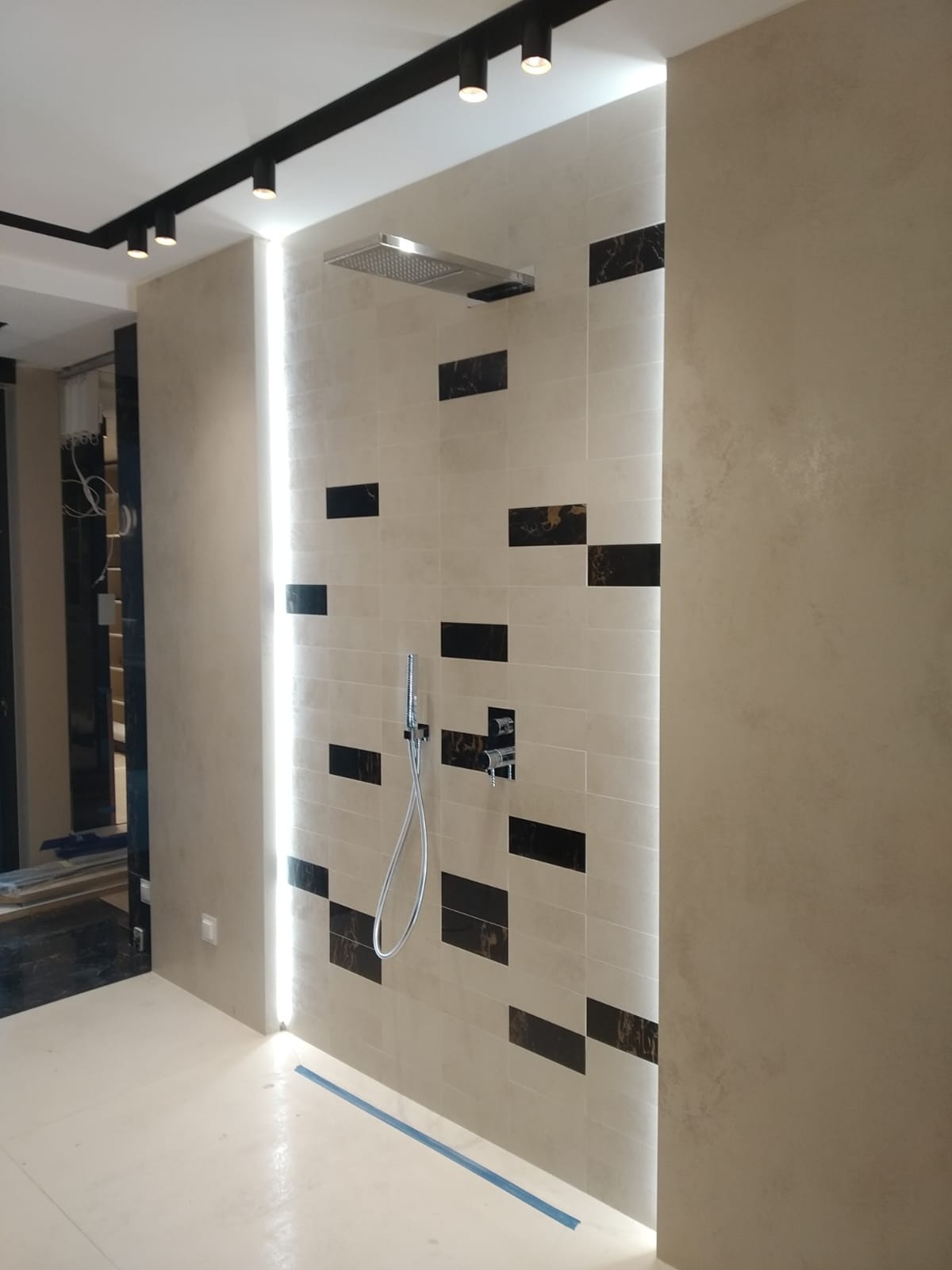 Wykończona kabina prysznicowa z beżowymi płytkami ułożonymi w cegiełkę, urozmaiconymi poziomymi wstawkami z płytek imitujących czarny marmur, oświetlona pionową listwą LED, z chromowaną...