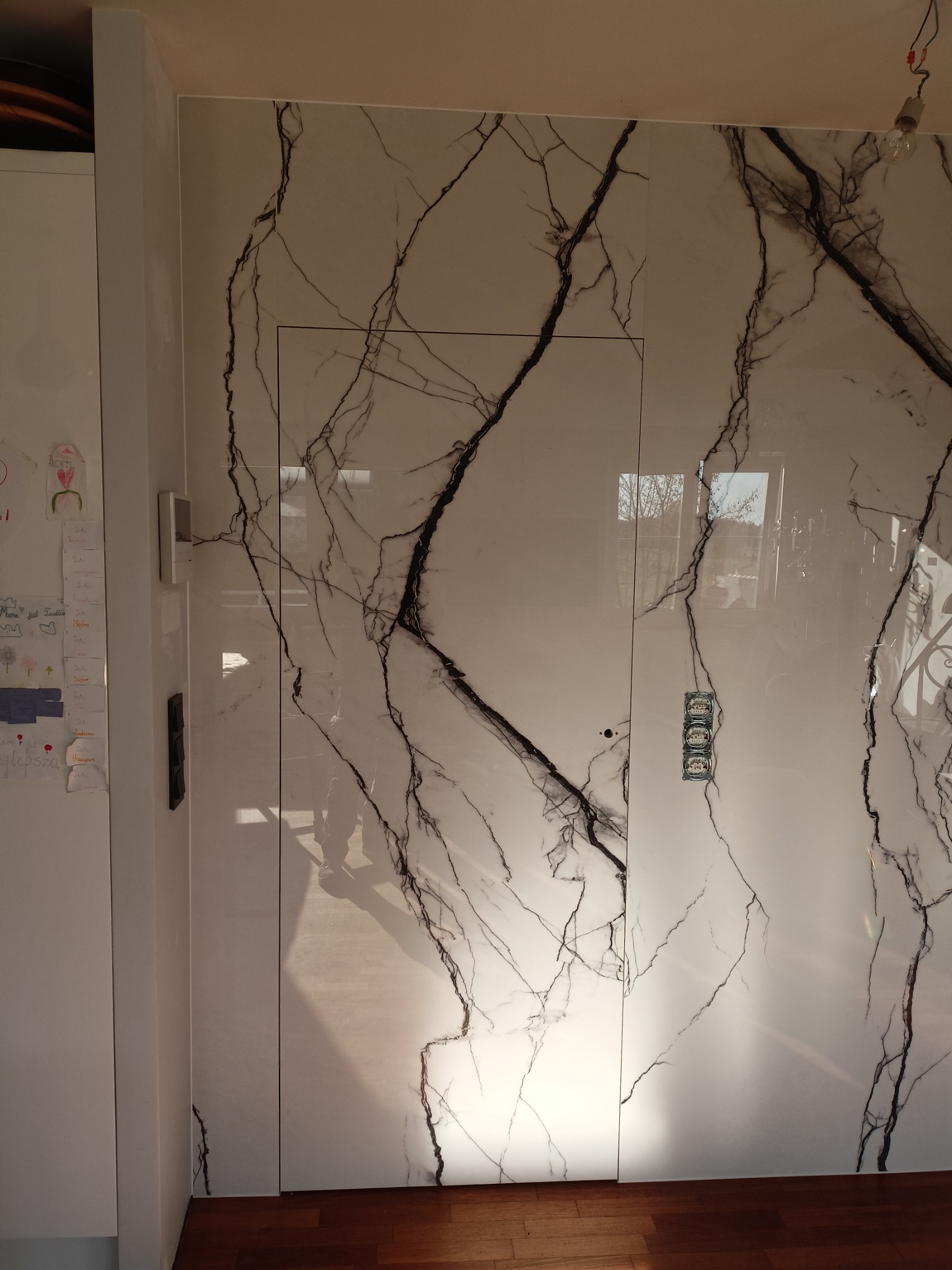 Ściana wyłożona dużymi płytami imitującymi biały marmur z czarnymi żyłami, ukryte drzwi wlicowane w ścianę, widoczne gniazdka elektryczne