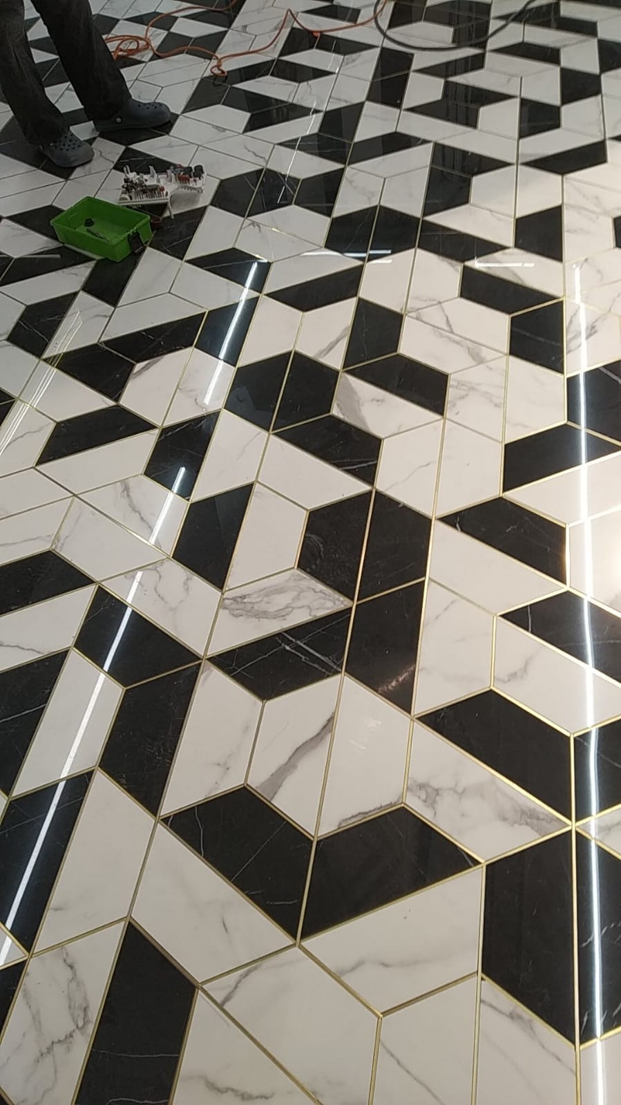 Ułożona podłoga z czarno-białych płytek imitujących marmur, z geometrycznym wzorem 3D i złotymi fugami, widoczne narzędzia i fragment osoby w rogu kadru.