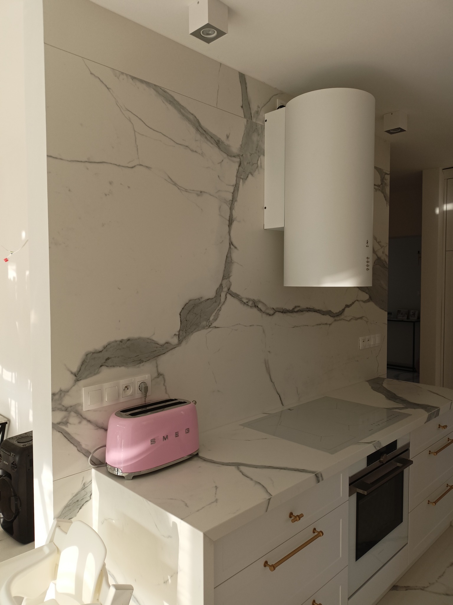 Minimalistyczna kuchnia z białymi szafkami, blatem i ścianą z płytek imitujących marmur, różowy toster SMEG i biały okrągły okap.