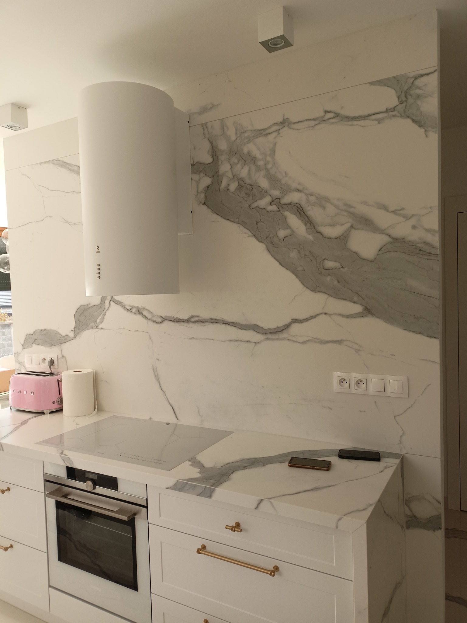 Biała kuchnia z marmurowym blatem i ścianą, nowoczesny okrągły okap, różowy toster SMEG i dwa smartfony na blacie.