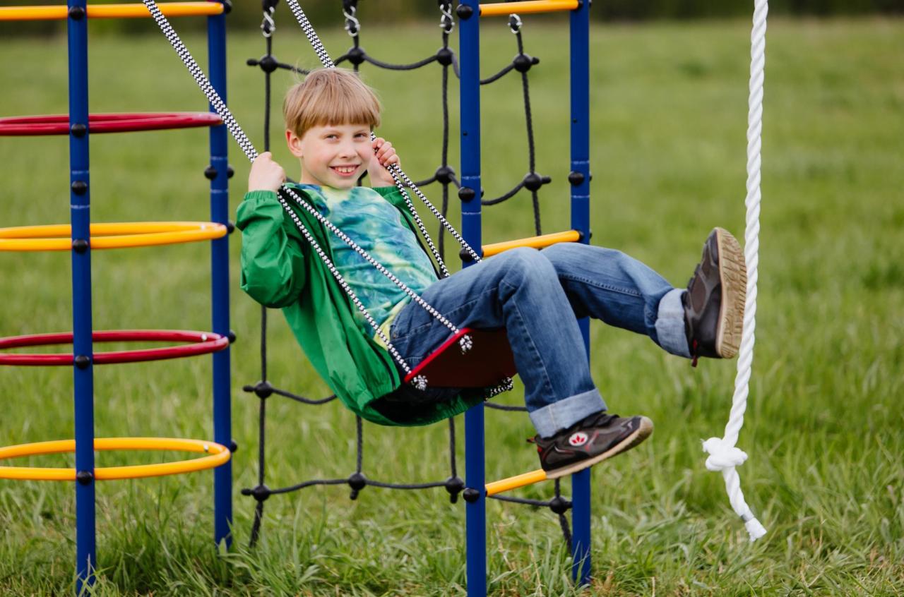 Uśmiechnięty chłopiec w zielonej kurtce huśta się na huśtawce linowej z czerwonym siedziskiem na tle zielonego trawnika i konstrukcji zabawowej z kolorowymi obręczami.
