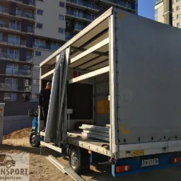 Otwarta skrzynia ładunkowa busy z częściowo zasuniętą plandeką, widoczny mężczyzna w środku i elementy ładunku, w tle budynek mieszkalny.