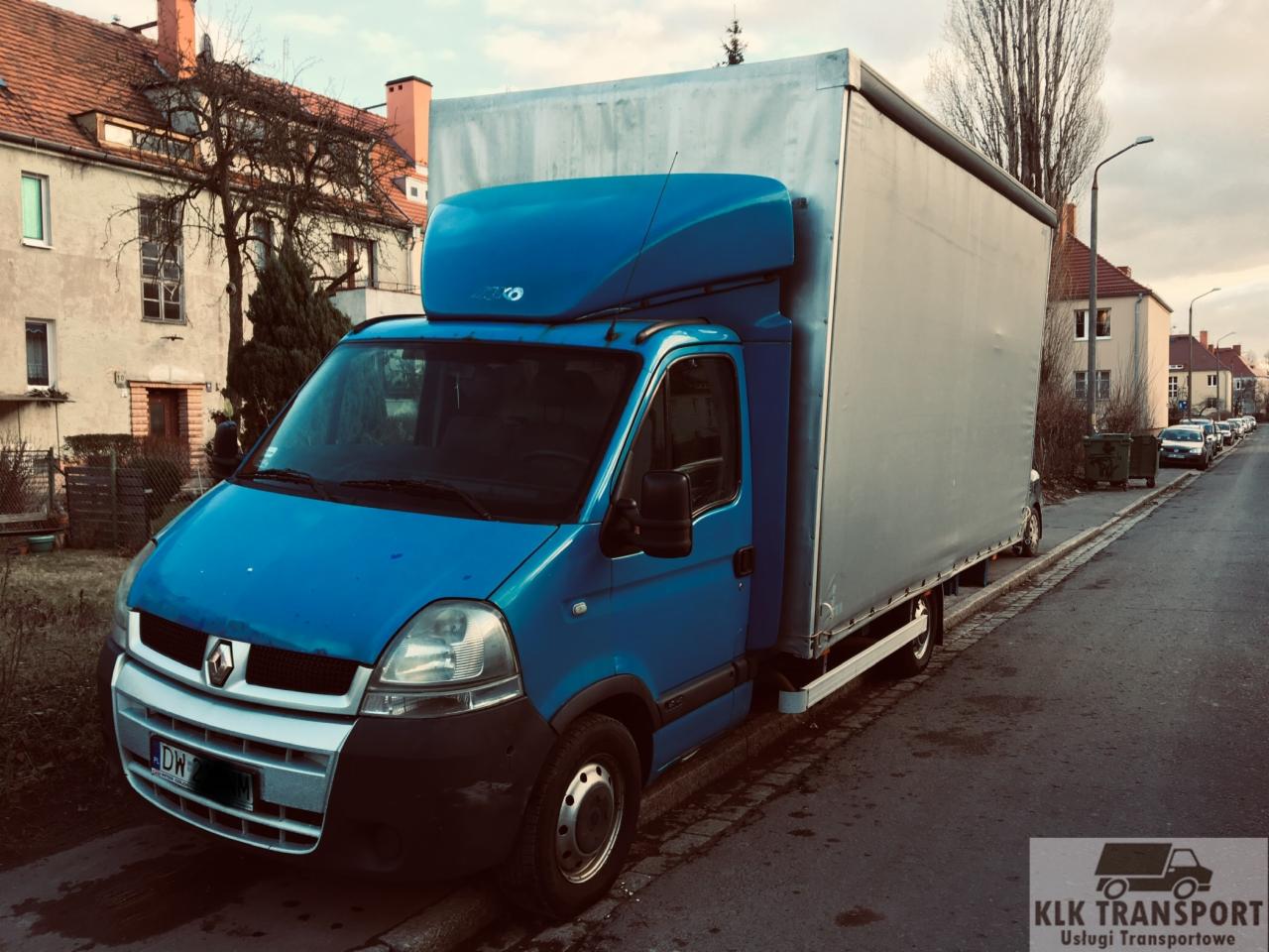 Niebieski samochód dostawczy Renault Master z plandeką na tle ulicy miejskiej i budynków mieszkalnych. Widoczne logo firmy transportowej w prawym dolnym rogu.