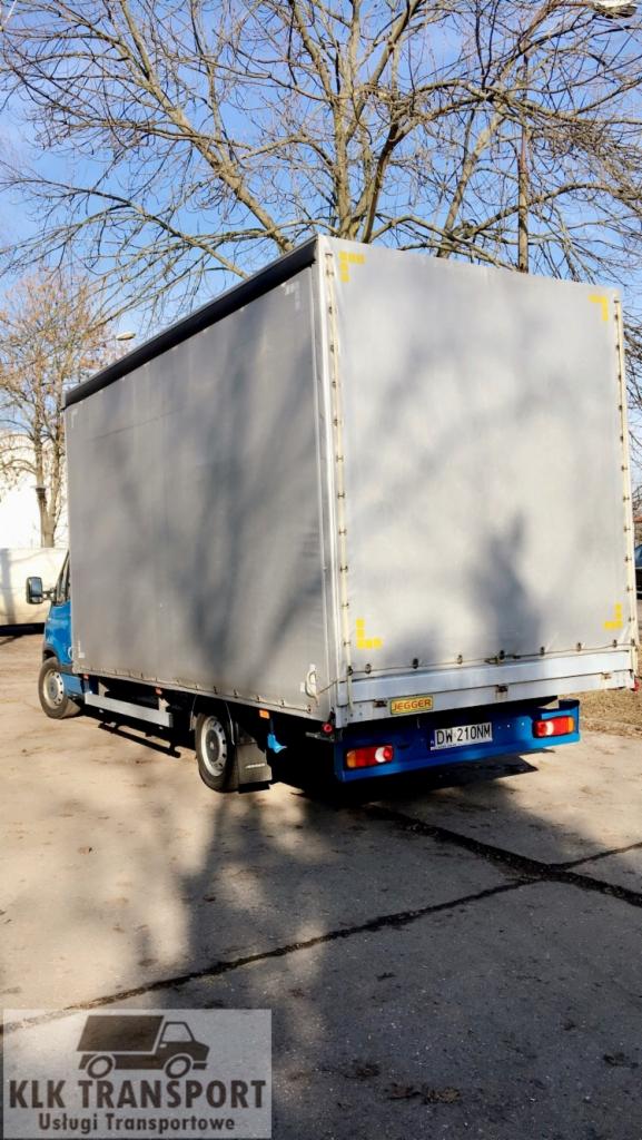 Niebieski samochód dostawczy z plandeką na parkingu, widok z tyłu, tablica rejestracyjna DW 210NM, z logo firmy transportowej w rogu.