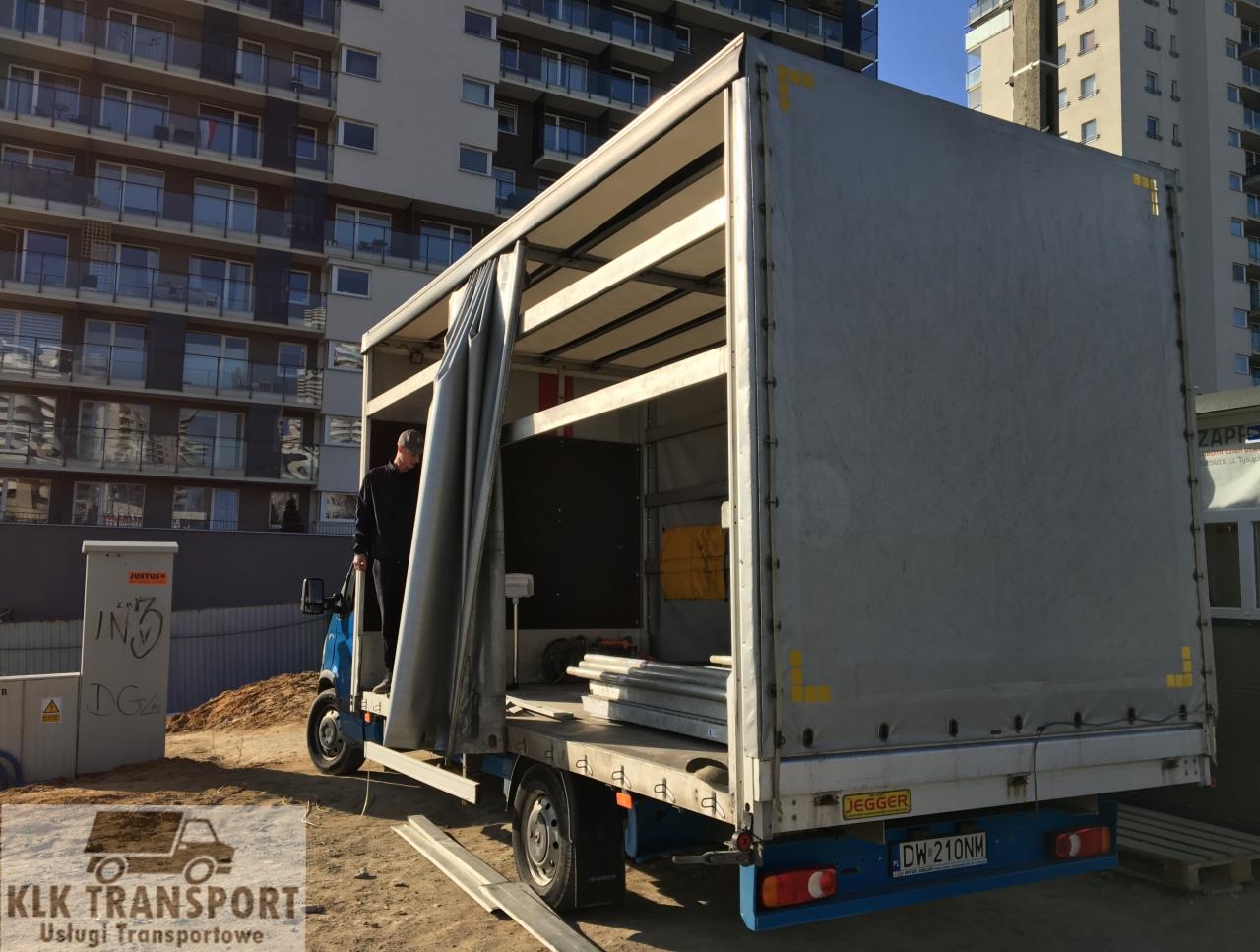 Otwarta skrzynia ładunkowa busy z częściowo zasuniętą plandeką, widoczny mężczyzna w środku i elementy ładunku, w tle budynek mieszkalny.