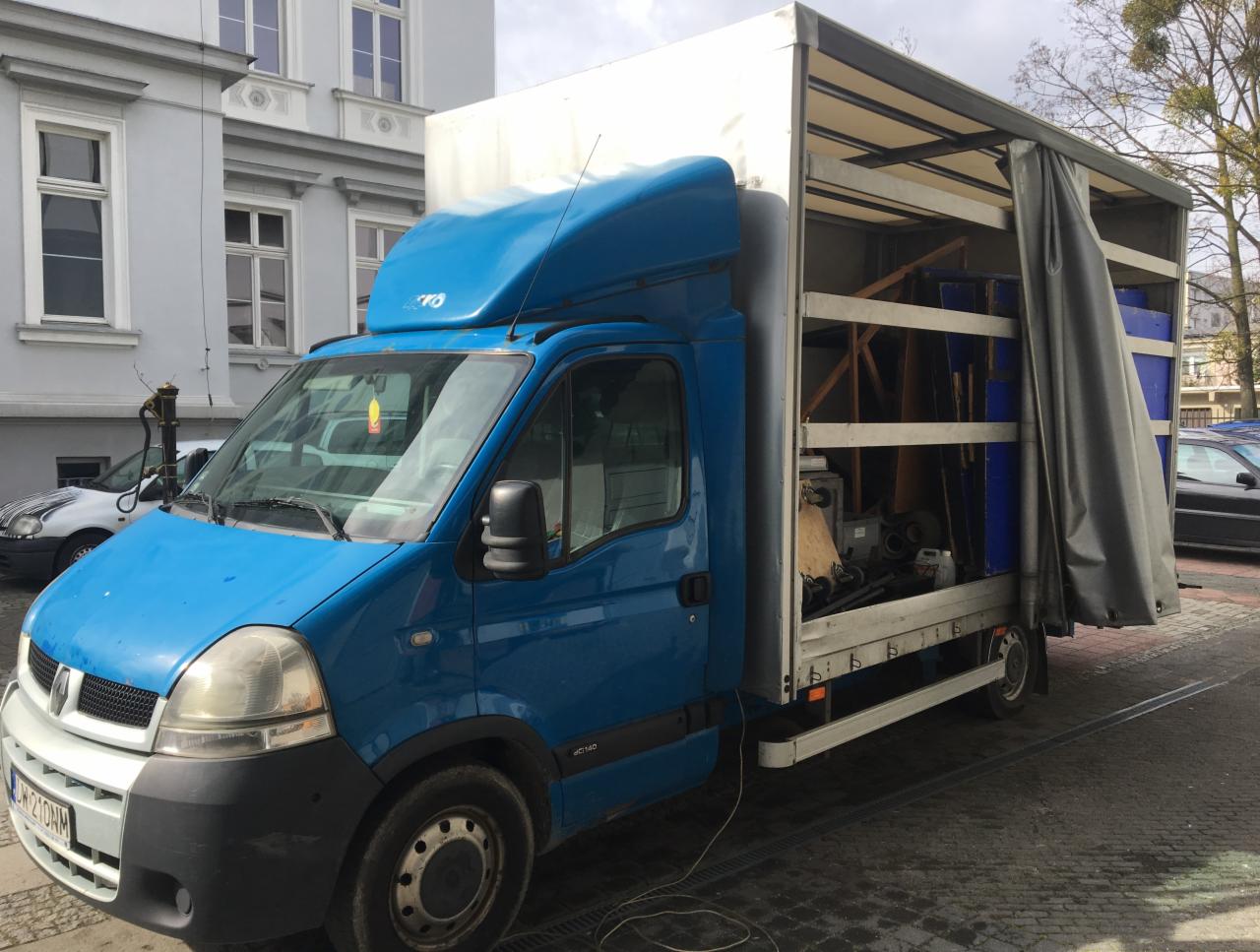 Niebieski bus Renault Master z otwartą przestrzenią ładunkową, częściowo zasłoniętą szarą plandeką, wypełnioną meblami i innymi przedmiotami podczas przeprowadzki na tle miejskiej zabudowy.