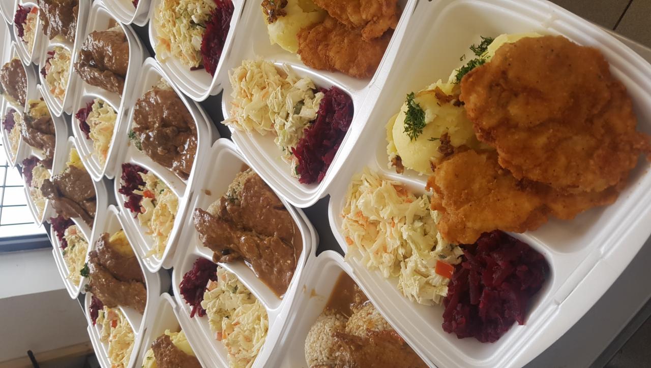 Rzędy gotowych dań obiadowych w białych, plastikowych pojemnikach cateringowych: kotlet schabowy z ziemniakami i surówkami oraz mięso w sosie z kaszą gryczaną i surówkami.