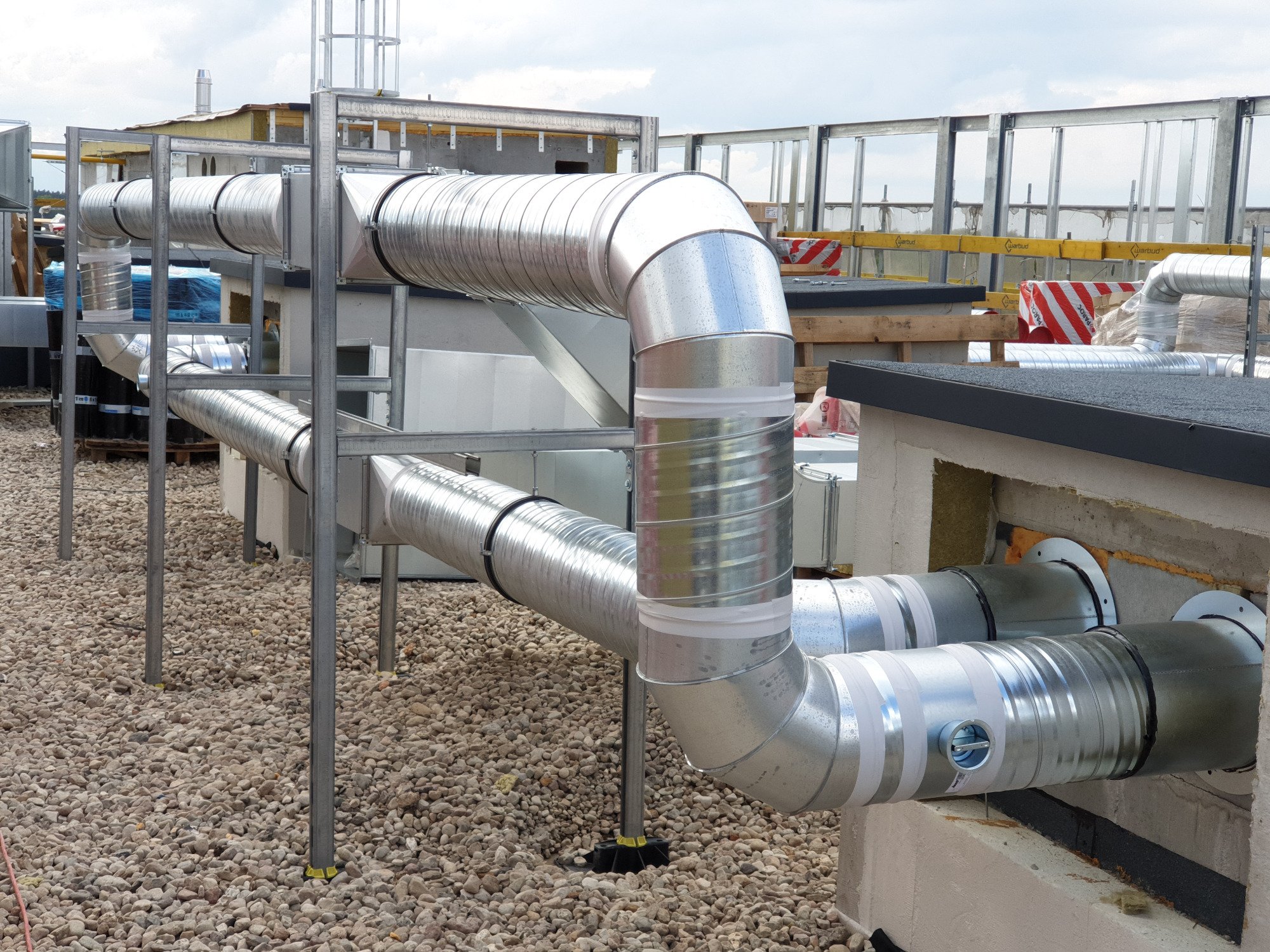 System kanałów wentylacyjnych z ocynkowanej blachy, zamontowany na dachu budynku pokrytym żwirem. Widoczne elementy mocujące i izolacja na łączeniach. Konstrukcja wsporcza z metalowych profili.