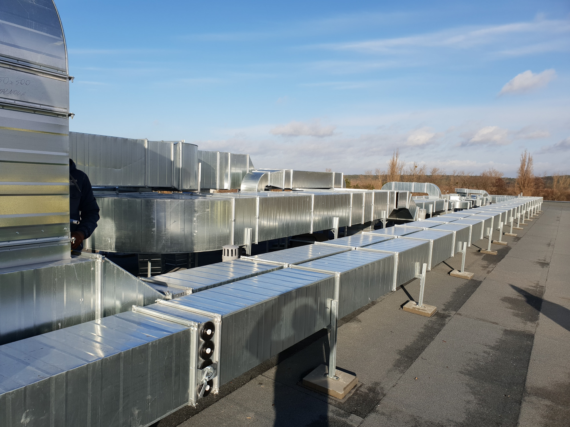 Długi szereg prostokątnych kanałów wentylacyjnych z ocynkowanej blachy, zamontowanych na dachu płaskim, wspartych na stalowych podporach z betonowymi podstawami, pod błękitnym niebem.