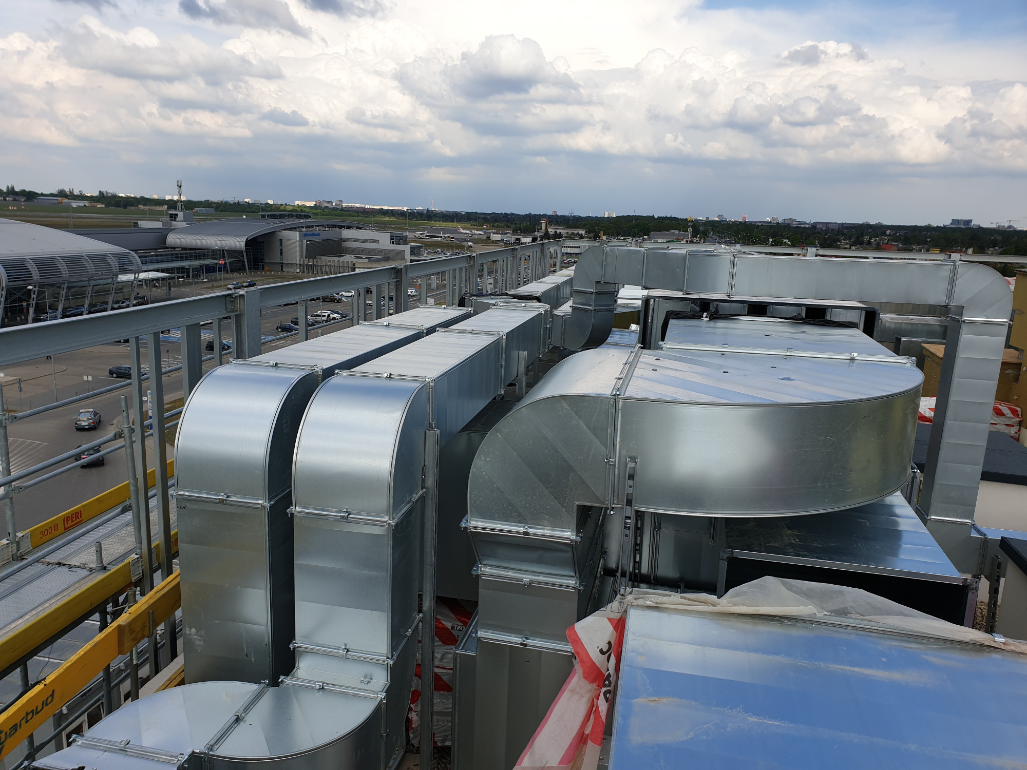 Widok z góry na system wentylacji z ocynkowanej stali na dachu budynku, w tle lotnisko z widocznymi pasami startowymi i terminalem, pochmurne niebo.