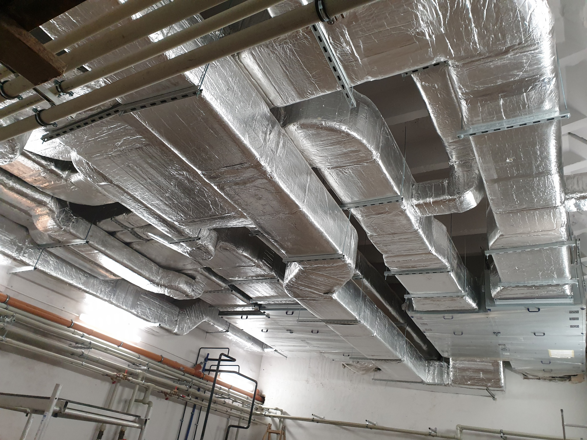 System kanałów wentylacyjnych z izolacją aluminiową, zamontowany pod sufitem, widoczne wsporniki mocujące i dodatkowe rury instalacyjne.