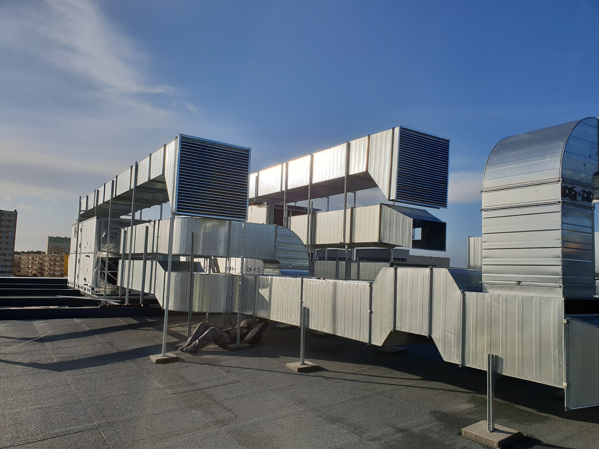 Instalacja kanałów wentylacyjnych na dachu budynku, widoczny pracownik w kombinezonie podczas montażu, system rur wentylacyjnych i kratki wentylacyjne, niebo w tle.